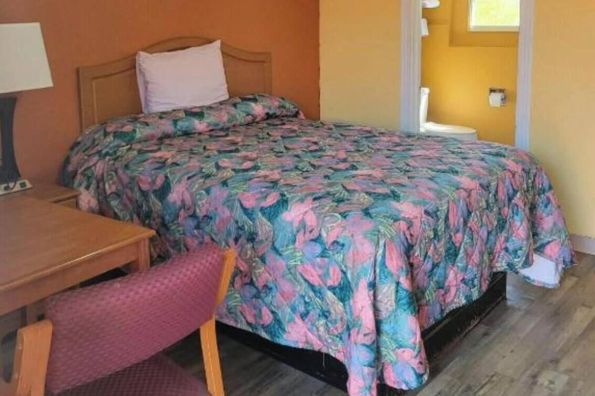 Una cama individual con una colcha estampada, una mesita de noche de madera, una silla y una ventana con cortinas.