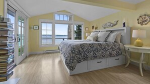 A bedroom with a bed, a bedside table, a lamp, a window with a view, and a stack of books. - Oceanfront Retreat on Spruce Point, Private Dock, Hot Tub & Resort Amenities (Boothbay Harbor)