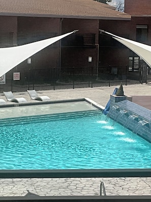 Una piscina con cascada, rodeada de sillas de descanso y un edificio de ladrillo al fondo.