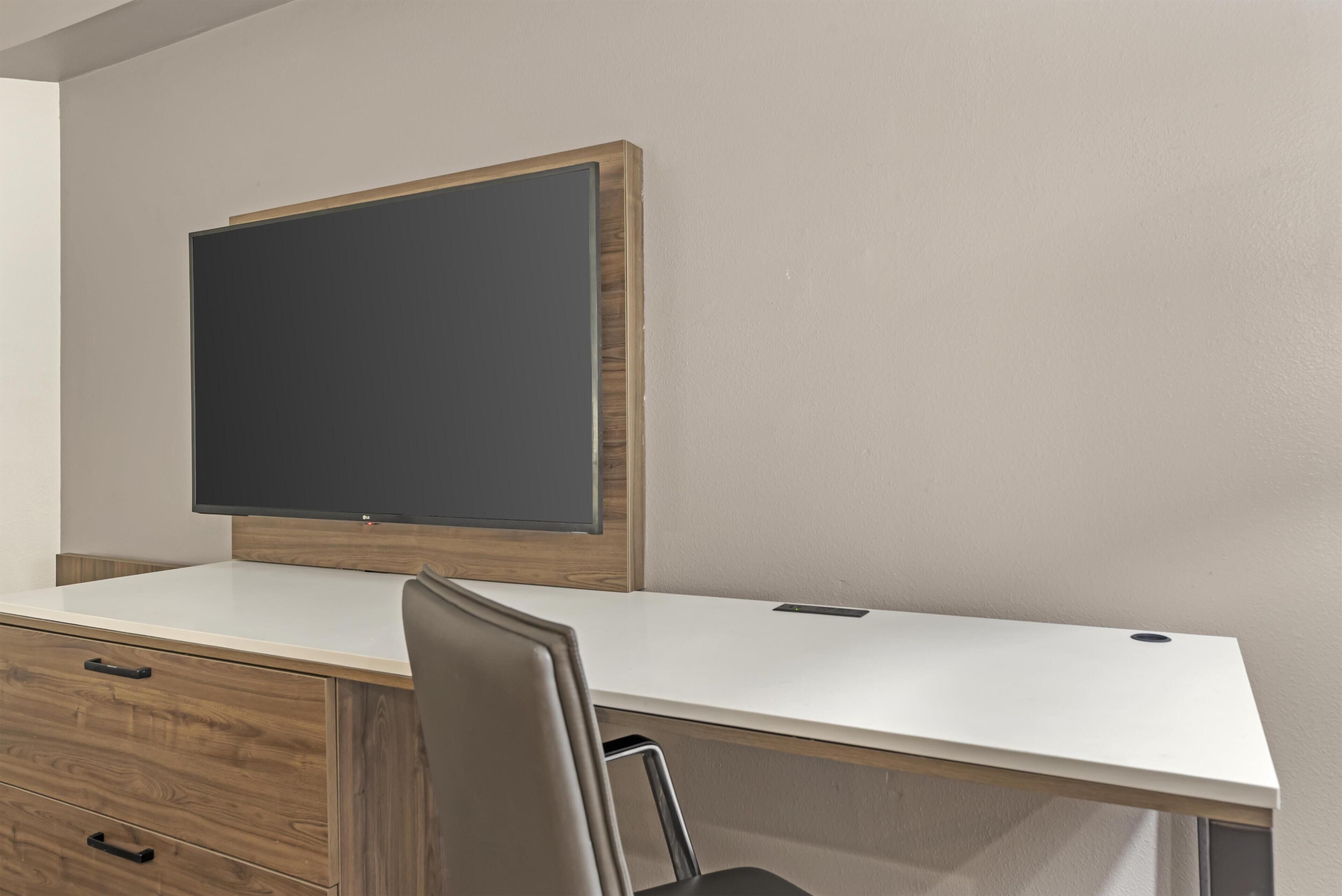 Una habitación moderna con un escritorio blanco, una silla, un mueble de madera y un televisor de pantalla plana grande montado en la pared.