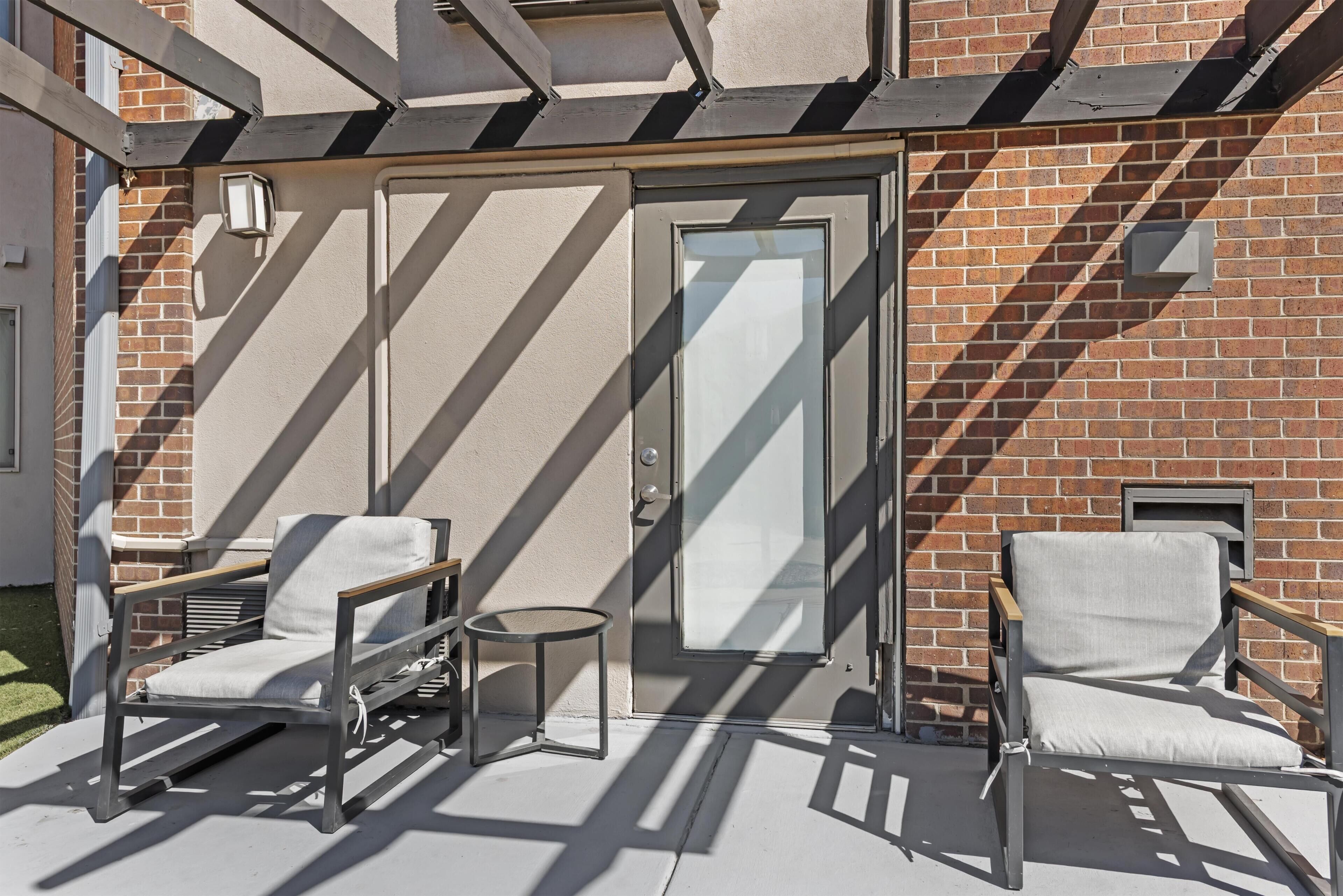 A patio with two chairs, a small round table, and a glass door leading to a brick building.