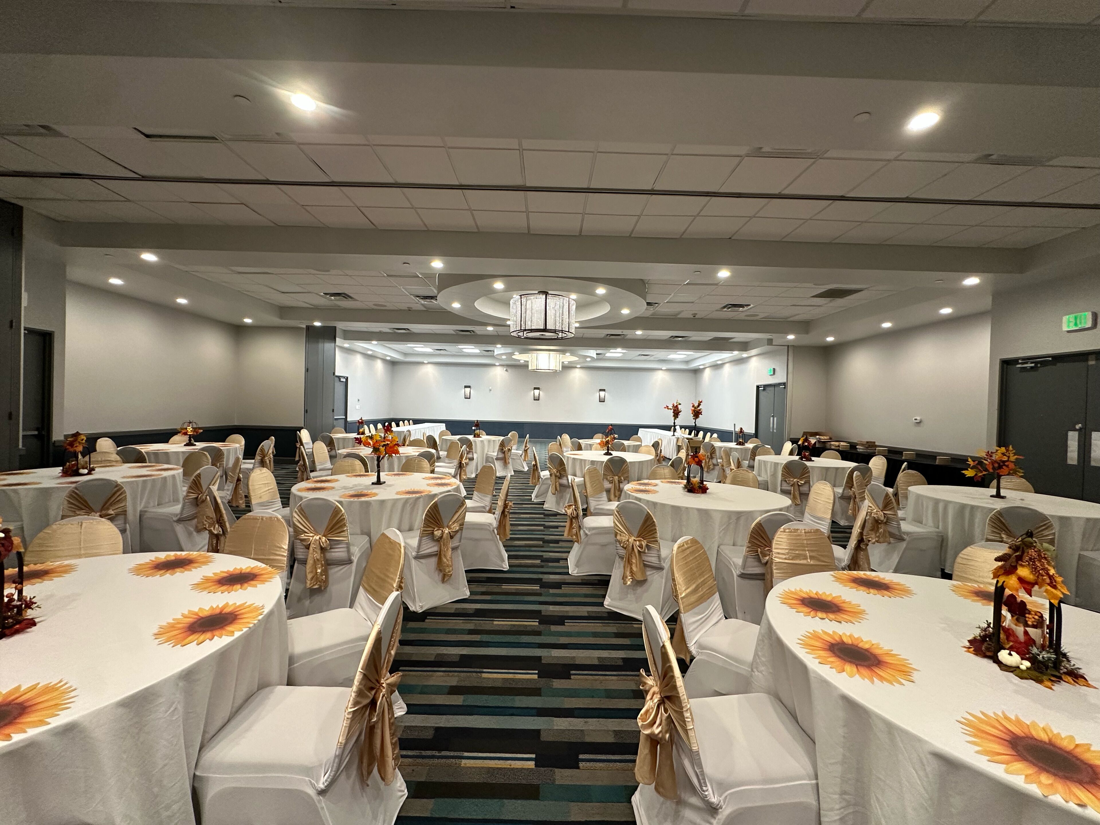 Une salle de conférence dotée de plusieurs tables rondes, chacune recouverte d’une nappe blanche et agrémentée de tournesols, ainsi que de chaises ornées de rubans beige et doré.