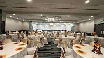 Une salle de conférence dotée de plusieurs tables rondes, chacune recouverte d’une nappe blanche et agrémentée de tournesols, ainsi que de chaises ornées de rubans beige et doré.