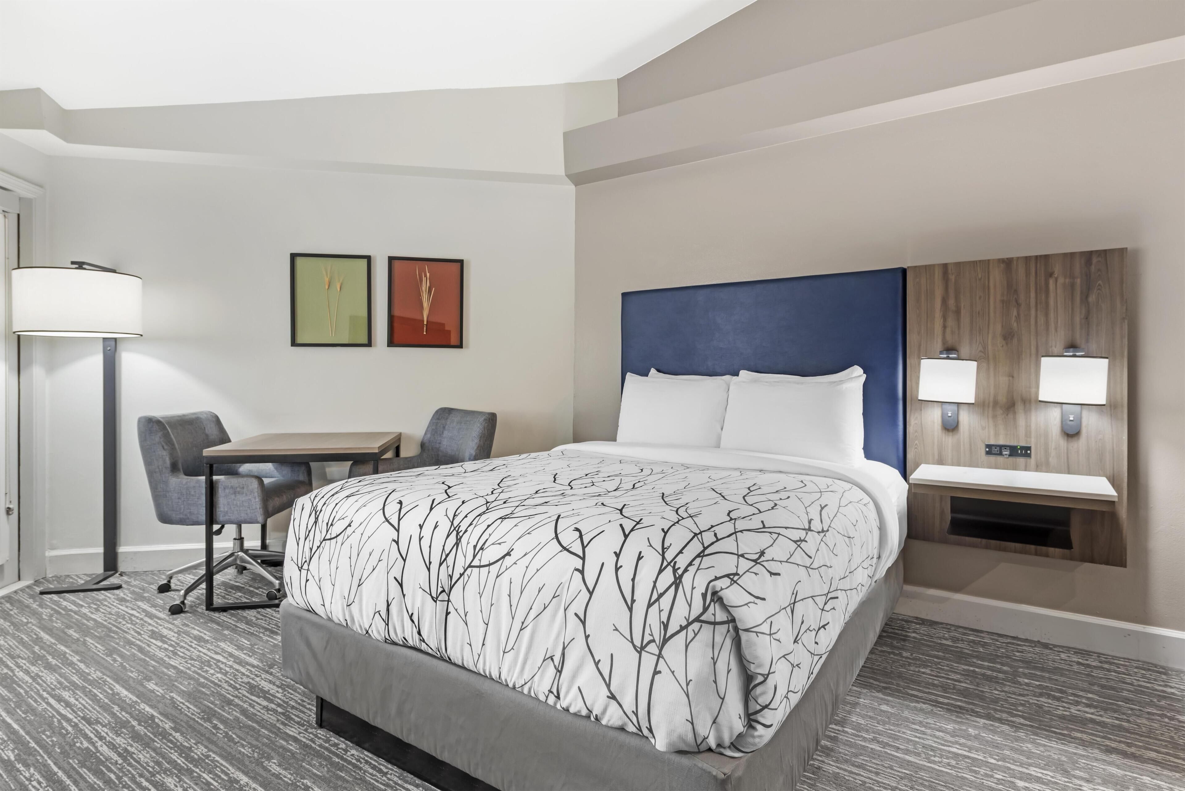 A hotel room with a bed, desk, chair, lamp, and two wall-mounted lights.
