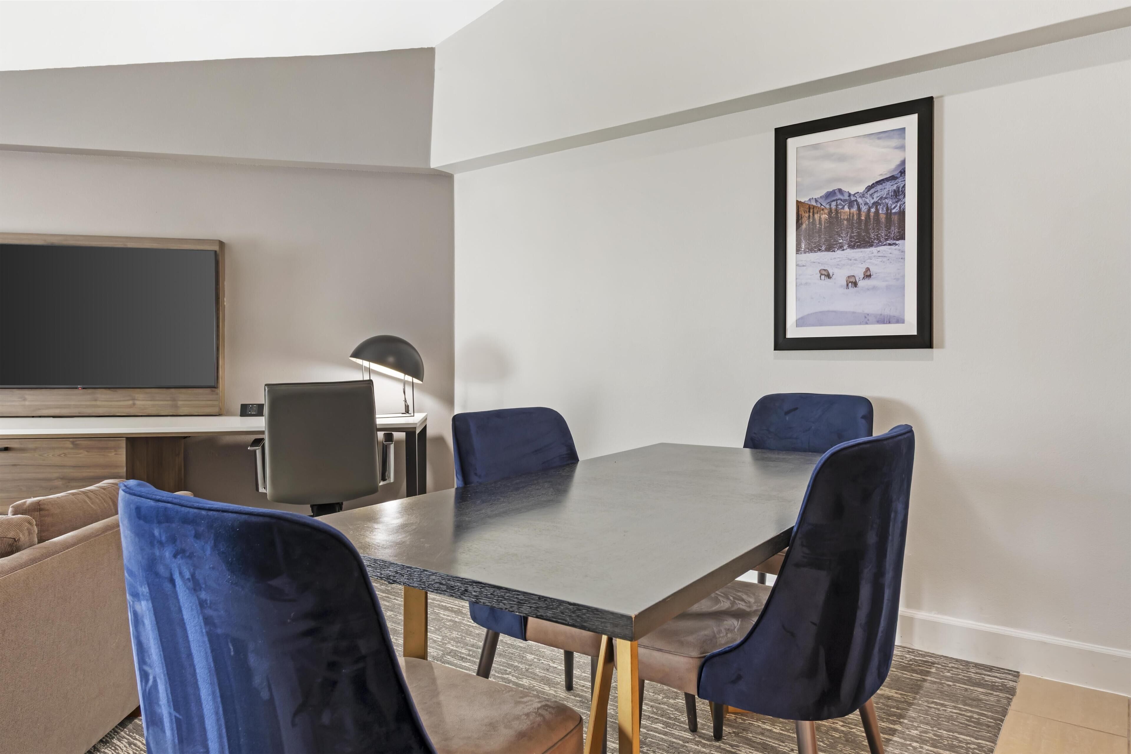 A modern dining area with a rectangular table, four blue chairs, a television, and a framed picture on the wall.