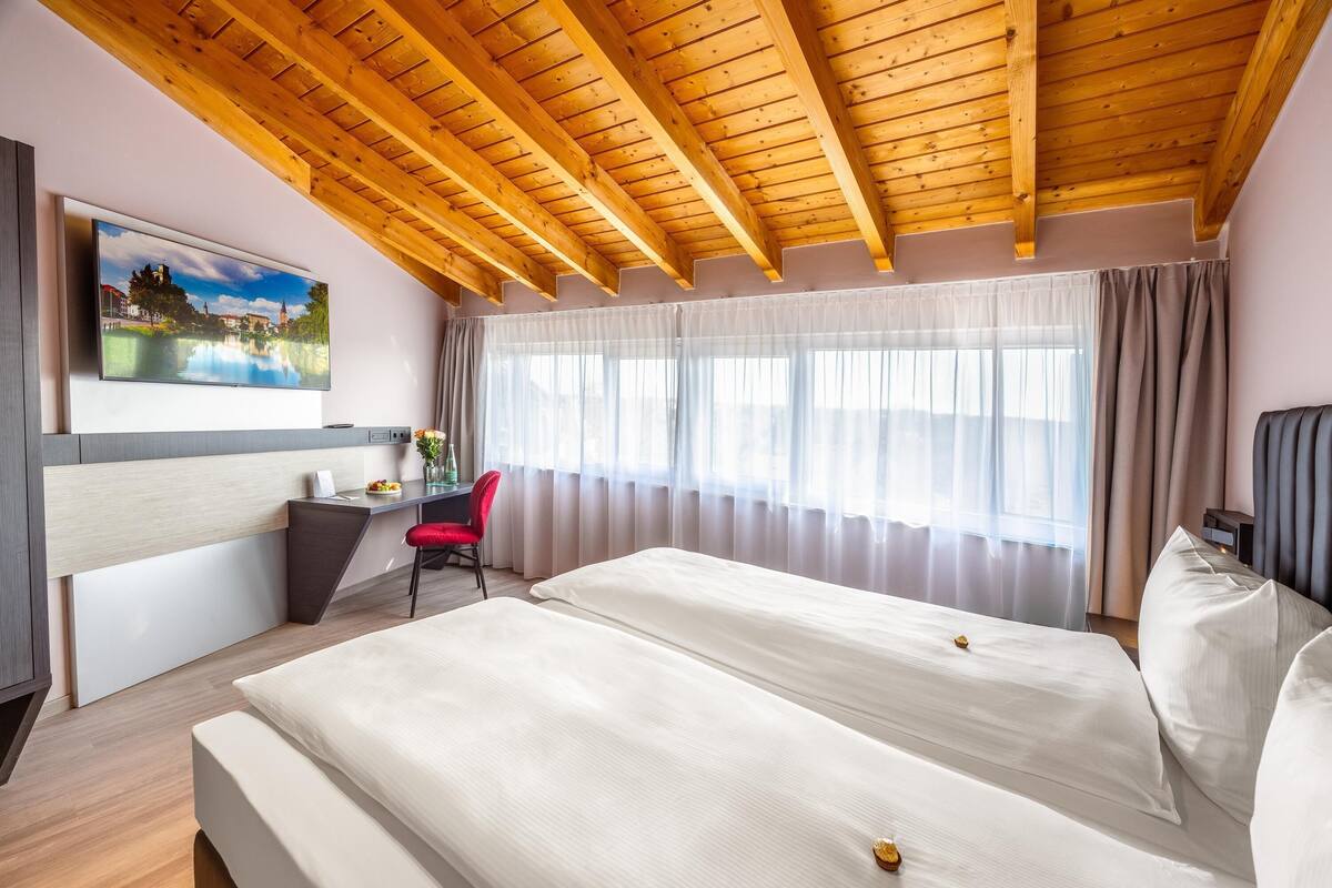 A hotel room with a wooden ceiling, a bed, a desk, a chair, and a window with curtains.
