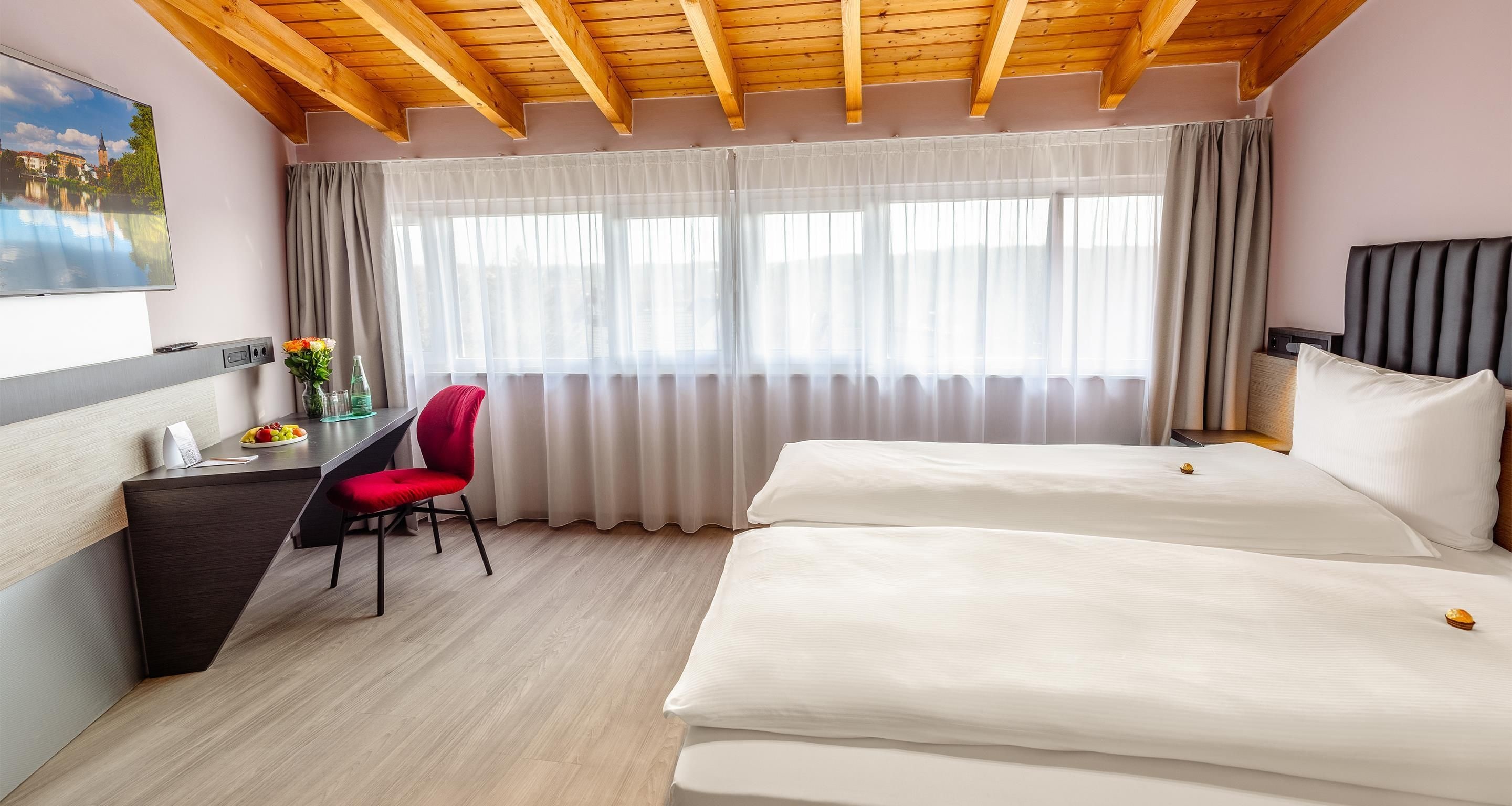 Habitación de hotel con techo de madera, un escritorio con silla, cama y un ventanal con cortinas.