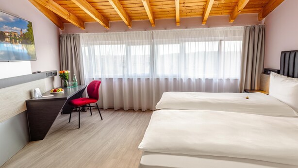 Habitación de hotel con techo de madera, un escritorio con silla, cama y un ventanal con cortinas.