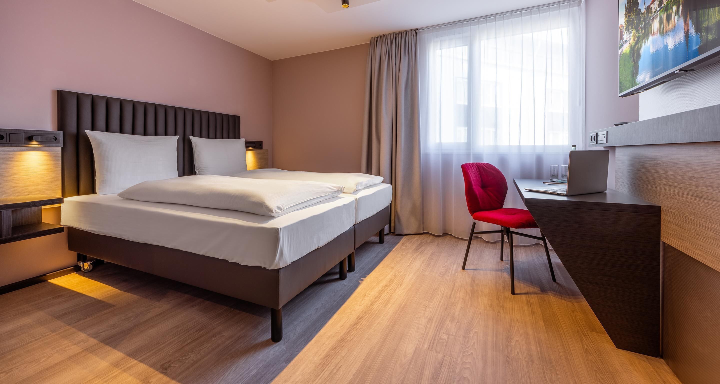 Quarto de hotel com cama, escrivaninha, cadeira, televisão e janela com cortinas.