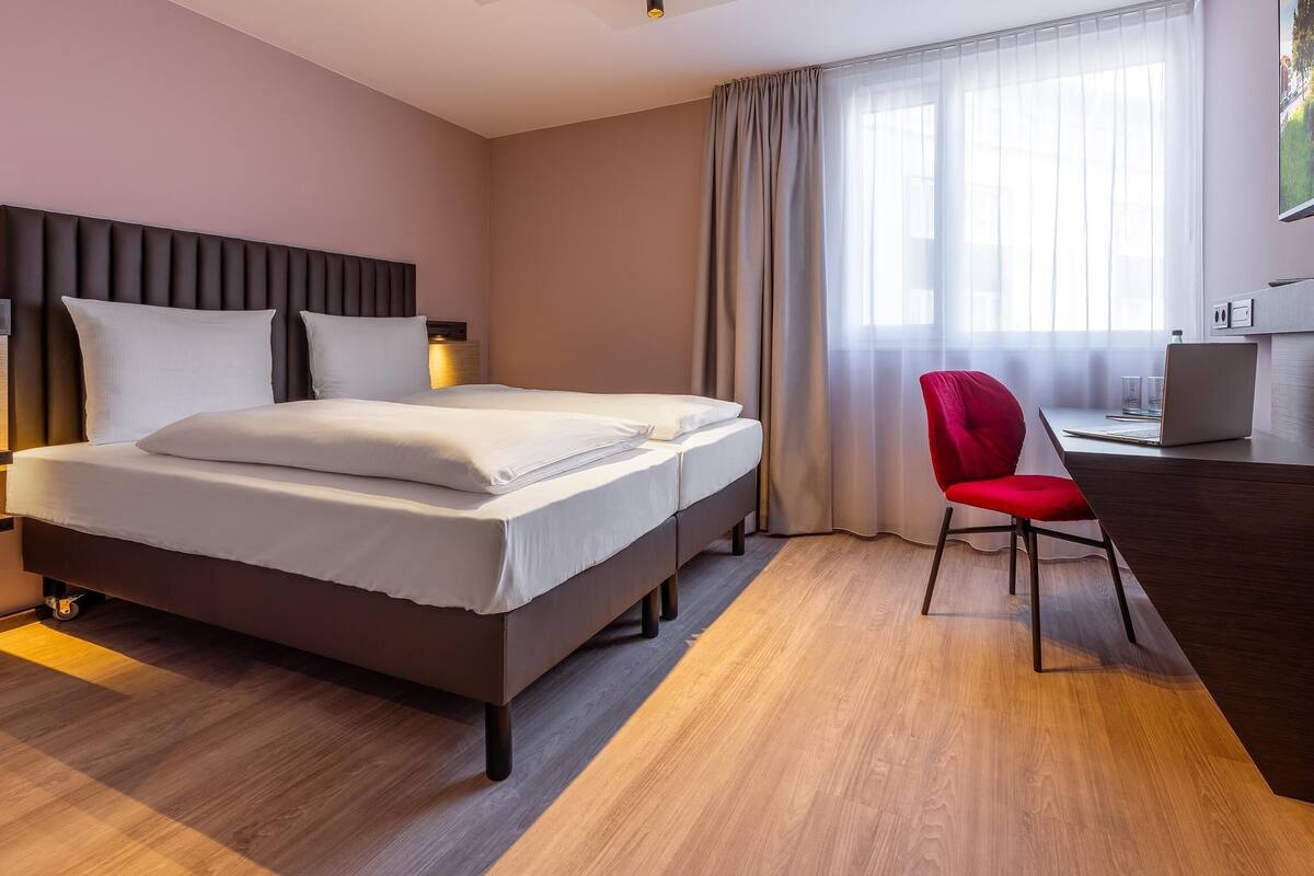 A hotel room with a bed, a desk, a chair, a TV, and a window with curtains.