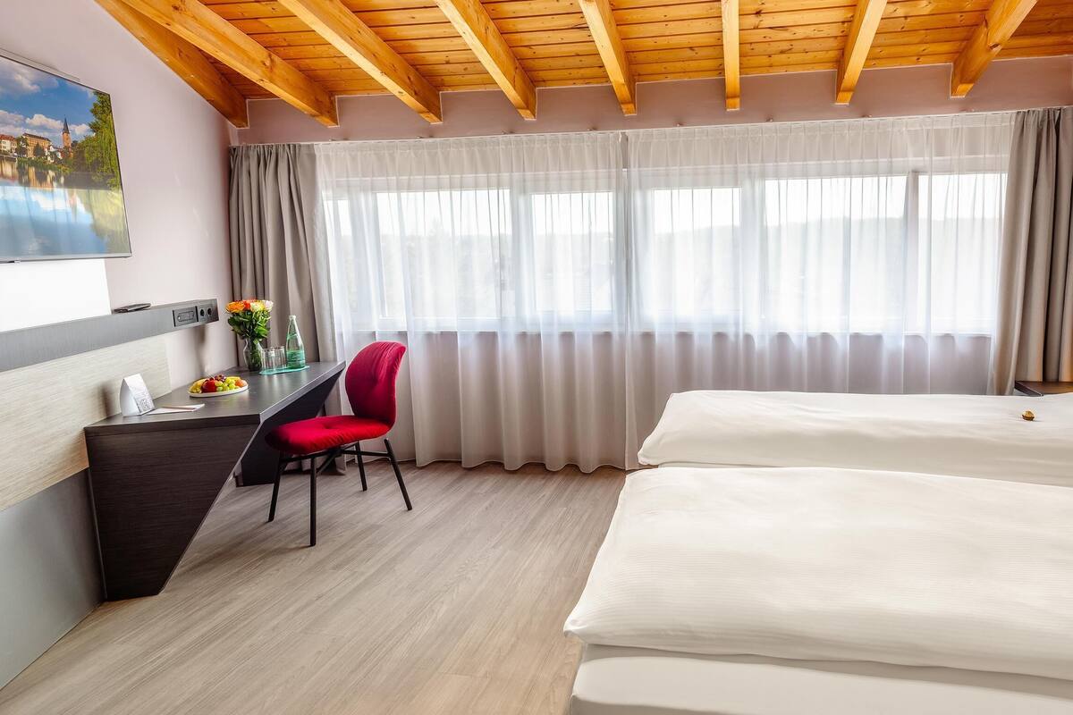 A hotel room with a wooden ceiling, a desk with a chair, two beds, and a large window with curtains.