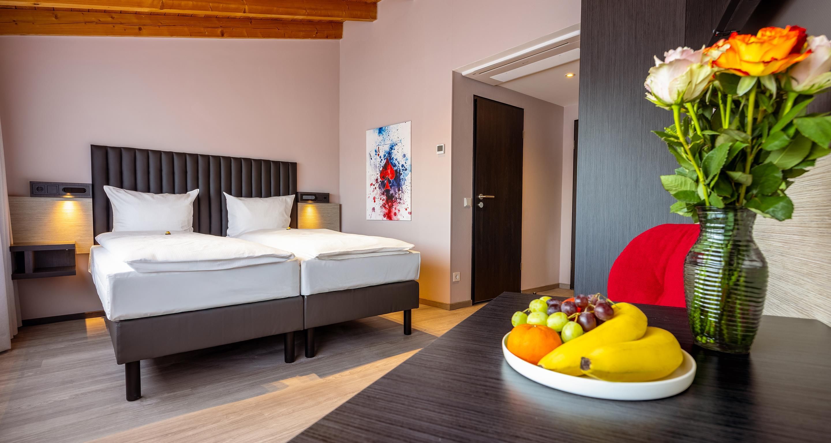 Ein Hotelzimmer mit einem Doppelbett, Nachttischen, einem Wandbild, einer Tür und einem Tisch mit Früchten.