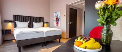 Quarto de hotel com cama de casal, mesas de cabeceira, uma obra de arte na parede, uma porta e uma mesa com frutas.