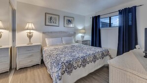 Un dormitorio con una cama, dos mesitas de noche, una lámpara, una ventana con cortinas y dos cuadros en la pared.