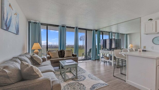 Amplia sala de estar con sofá, mesa de centro y televisor. Zona de comedor con sillas y mesa. La habitación tiene ventanas grandes con cortinas.