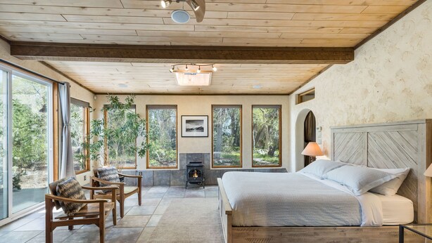 Ein Schlafzimmer mit einem großen Bett, Holzmöbeln, einem Kamin und großen Fenstern mit Blick auf Bäume.