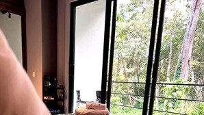 Una cama con sábana rosa, cabecera negra y vistas a árboles a través de amplias ventanas.