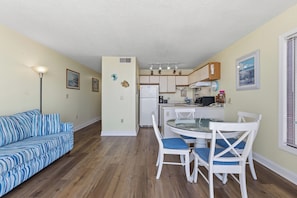 A living room with a sofa, a dining table with chairs, a kitchen area, and a ceiling fan. - Oceanfront 1BR condo in Carolina Beach with stunning views for 6 guests. (Carolina Beach)