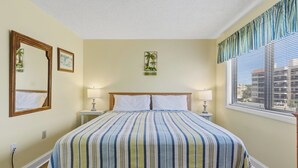 Ein Hotelzimmer mit einem Bett, zwei Nachttischen, einem Fenster mit Jalousien, einem Wandspiegel und einem gerahmten Bild.