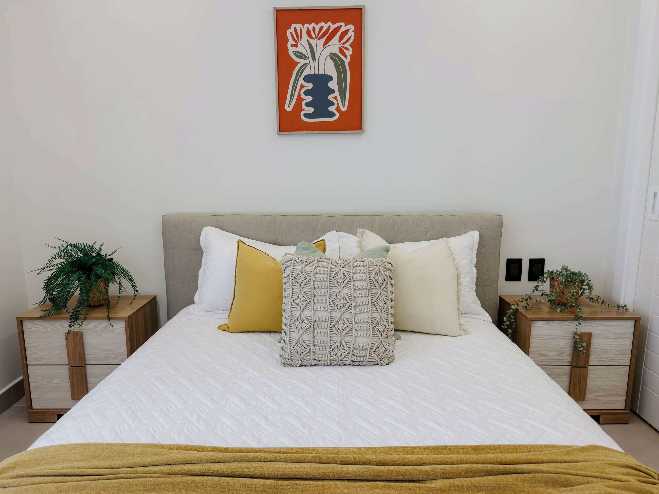 A neatly made bed with a white duvet, a mustard-colored throw blanket, and a patterned throw pillow. There are two nightstands with decorative plants, a framed artwork above the bed, and a light-colored wall.