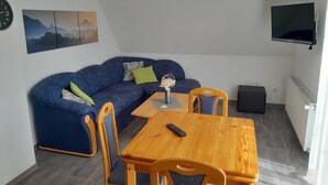 A living area with a blue sofa, wooden table, and chairs, a clock, and a mounted TV. - NEW! "Nordseeluft" Vacation Rental (Dornum)