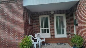 A brick building with two white doors, a covered patio area with two white plastic chairs, and two potted plants. - NEW! "Nordseeluft" Vacation Rental (Dornum)