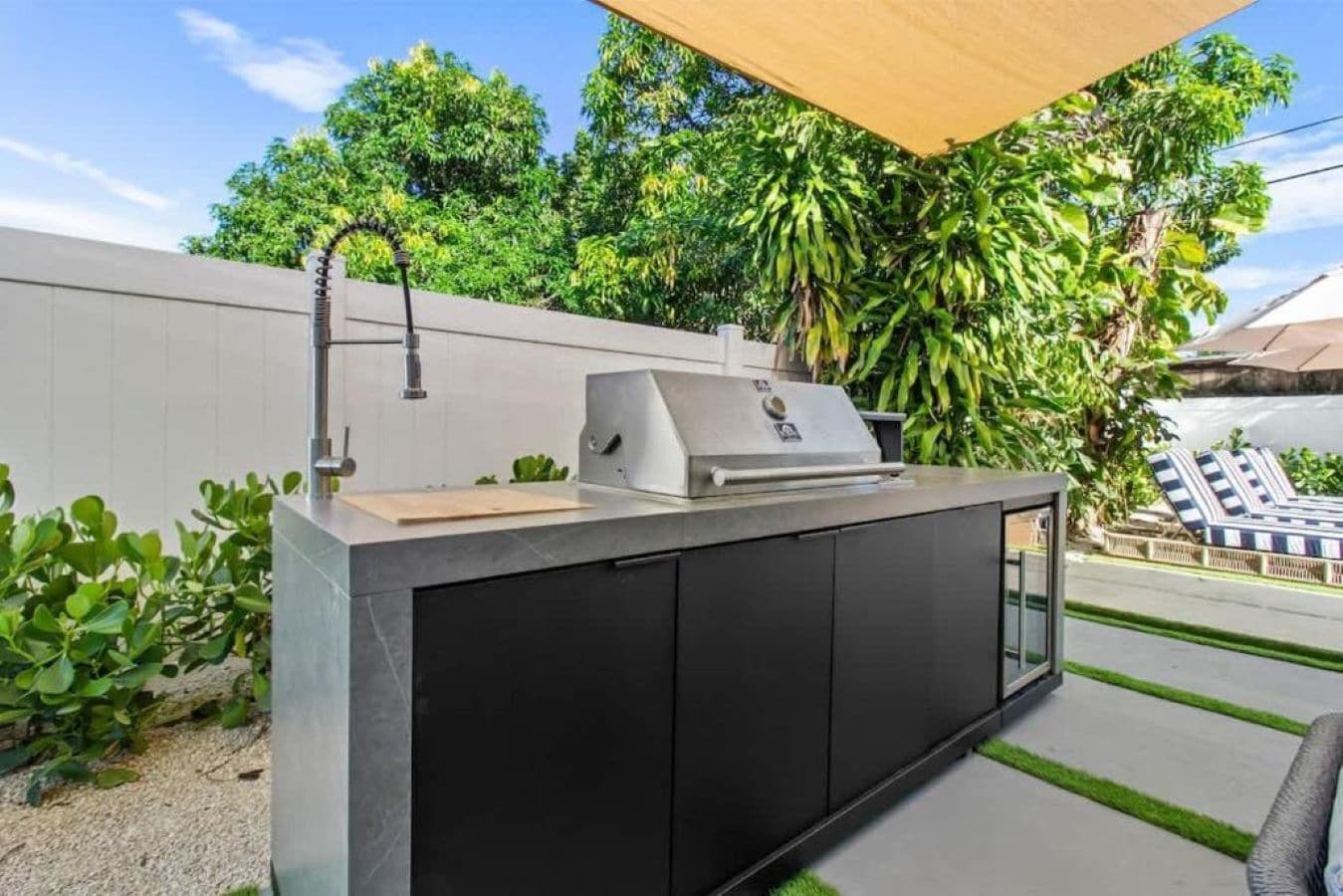 An outdoor kitchen with a grill, sink, and storage cabinets, surrounded by greenery and a pool area.