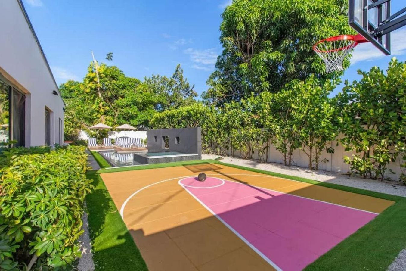 A basketball court with a hoop, surrounded by greenery and a building.
