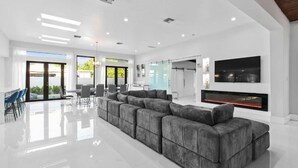 A spacious living area with a large grey sectional sofa, a flat-screen TV mounted on the wall, and a fireplace. - Luxury Miami Stay by Glam Homes (North Miami Beach)