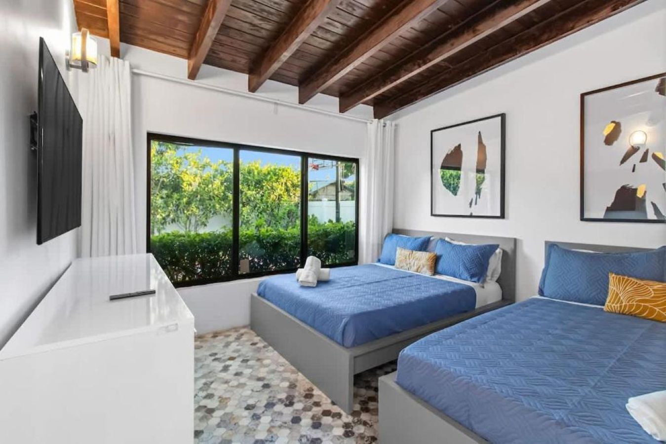 A modern hotel room with two beds, a TV, and large windows with a view of greenery.