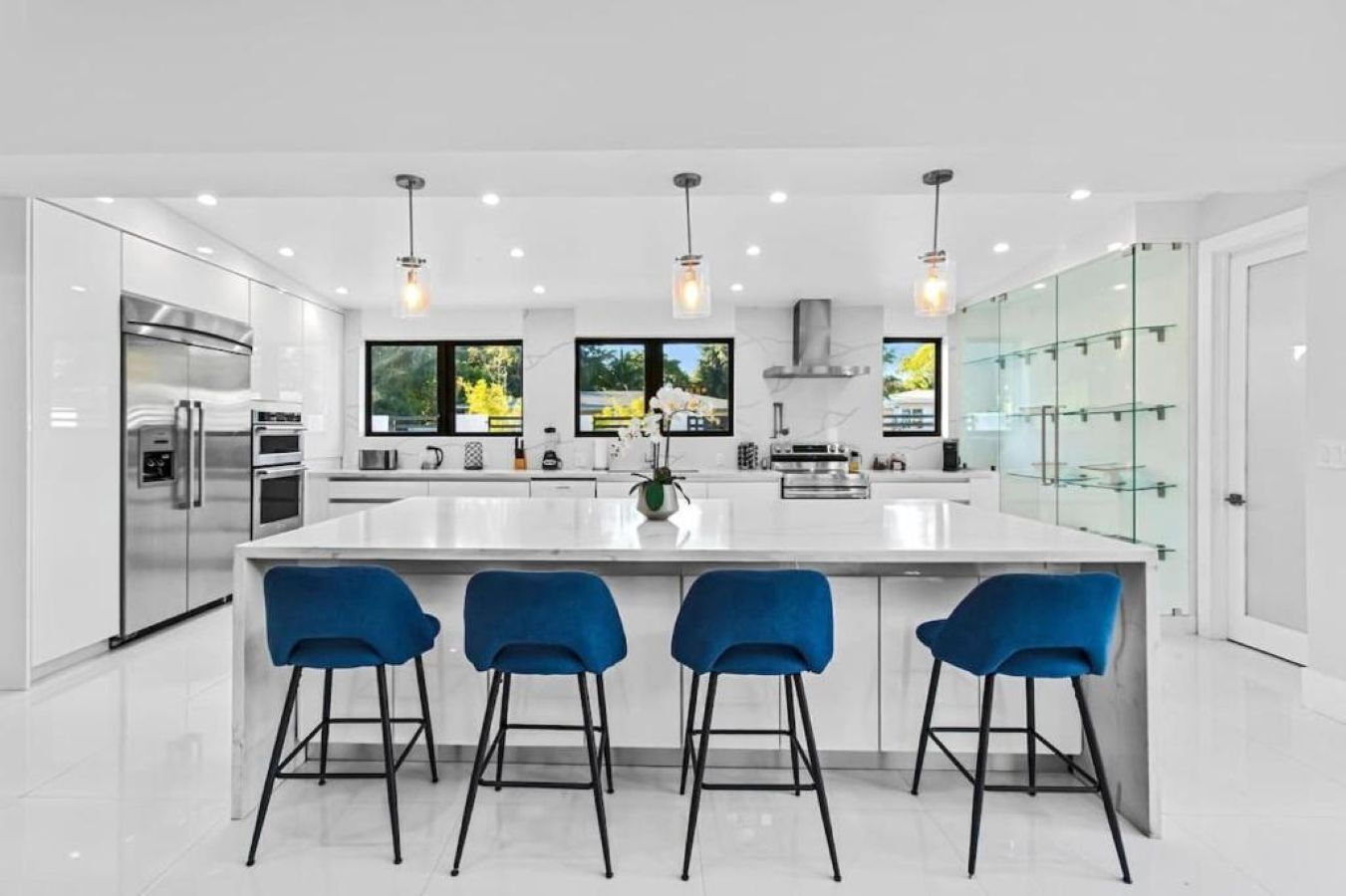 Een moderne keuken met een groot eiland, barstoelen, roestvrijstalen apparatuur en grote ramen.