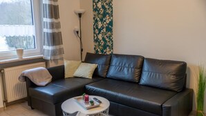 A black leather sofa with cushions, a small round coffee table, a radiator, a window with curtains, and a floral wallpaper.