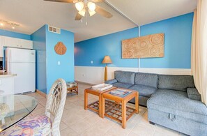 Un salon comprenant un canapé, une table basse en verre, un fauteuil en osier, un ventilateur de plafond et un coin cuisine avec un réfrigérateur.