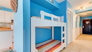 Dormitorio con literas, paredes azules, una zona de cocina con armarios blancos y una puerta que da a otra habitación.