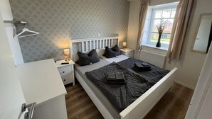 A bedroom with a bed, bedside tables, a window with curtains, a radiator, and a wall-mounted light fixture. - Vacation home Alte Schule (Dornumersiel)