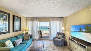 Ein Wohnzimmer mit Sofa, Sesseln, Fernseher und Blick auf den Strand durch eine Schiebetür.