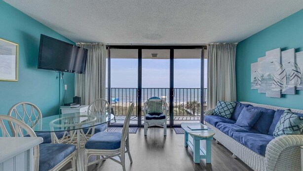 Ein Zimmer mit Balkon, einem Esstisch mit Stühlen, einer Couch und einem Fernseher an der Wand.