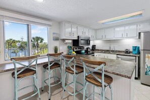 A kitchen with white cabinets, a microwave, a coffee maker, and a view of palm trees. - Pinnacle Port with Gulf & Lake Views | Pools | West End PCB (Panama City Beach)