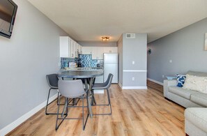 Eine moderne Küche mit weißen Schränken, ein Essbereich mit einem Glastisch und Metallstühlen sowie ein graues Sofa in einem Wohnzimmer.