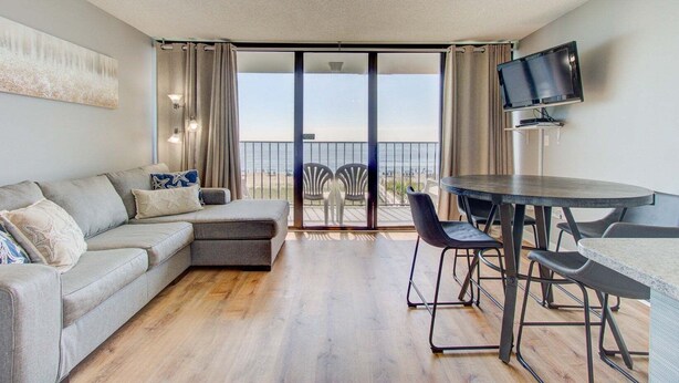 A modern living room with a grey sectional sofa, a dining table with chairs, a flat-screen TV, and a balcony with a view of the beach. - Experience oceanfront bliss at 0309 The Blue Depth in Carolina Beach (Carolina Beach)