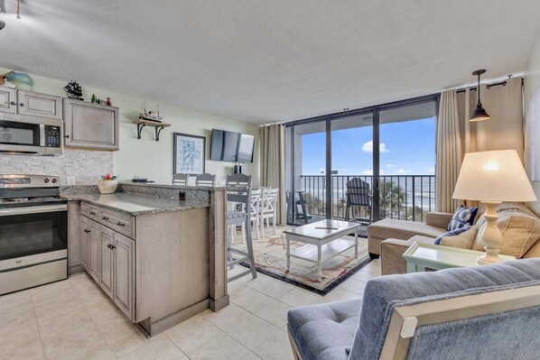 A modern kitchen with a large island, a dining area with white chairs, a sofa, and a balcony with a view of the ocean. - 0312 Captains Quarters: Stunning 2-bedroom oceanfront condo in Carolina Beach. (Carolina Beach)