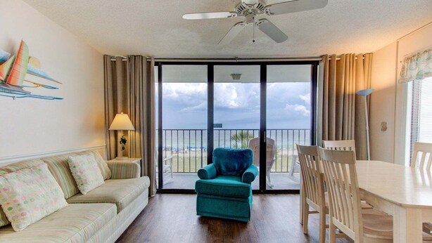 A room with a sofa, a teal armchair, a dining table with chairs, a ceiling fan, and a view of the ocean through large glass doors. - Charming 2BR oceanfront condo with stunning views at Carolina Beach 0314 (Carolina Beach)