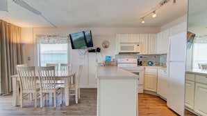 A modern kitchen with white cabinetry, a central island, a dining area with chairs, and a flat-screen TV mounted on the wall. - Charming 2BR oceanfront condo with stunning views at Carolina Beach 0314 (Carolina Beach)