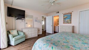 A bedroom with a bed, a television, a dresser, a chair, and a door leading to a bathroom. - Charming 2BR oceanfront condo with stunning views at Carolina Beach 0314 (Carolina Beach)