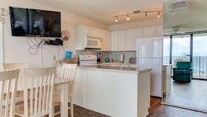 A modern kitchen with white cabinets, a dining area with wooden chairs, a flat-screen TV mounted on the wall, and a balcony with a view. - Charming 2BR oceanfront condo with stunning views at Carolina Beach 0314 (Carolina Beach)