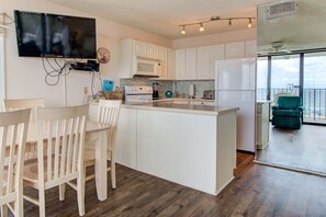 Eine moderne Küche mit weißen Schränken, ein Essbereich mit Holzsesseln, ein Flachbildfernseher an der Wand und ein Balkon mit Blick.