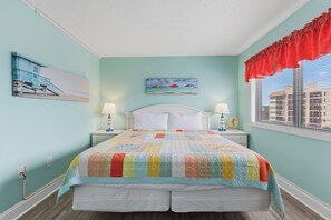 A bedroom with a bed, two bedside tables, a window with blinds, a red valance, and a beach-themed painting on the wall. - Experience beachfront bliss in this cozy 1-bedroom, Carolina Beach retreat. (Carolina Beach)
