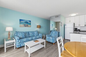 A living room with striped blue sofas, a white coffee table, and a kitchen area with white cabinets and a refrigerator. - Experience beachfront bliss in this cozy 1-bedroom, Carolina Beach retreat. (Carolina Beach)
