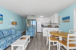 Eine moderne Küche mit weißen Schränken, ein Essbereich mit Holztisch und -stühlen sowie eine Wohnbereich mit gestreifter Couch und einer Lampe.