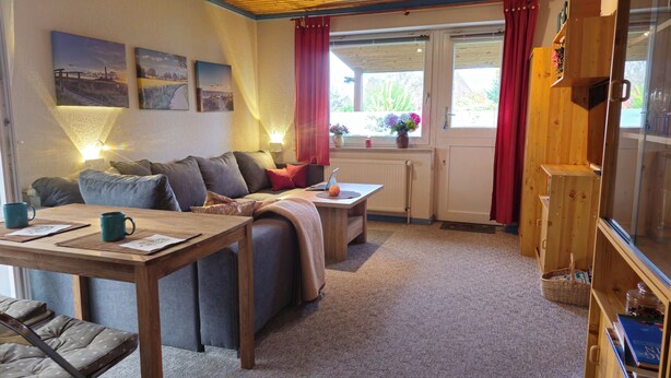 A cozy living room with a sofa, a wooden coffee table, and a dining table with two mugs. There are framed pictures on the wall, a window with red curtains, and a wooden cabinet. - Baltic Sea, Schlei and surroundings (Norderbrarup)